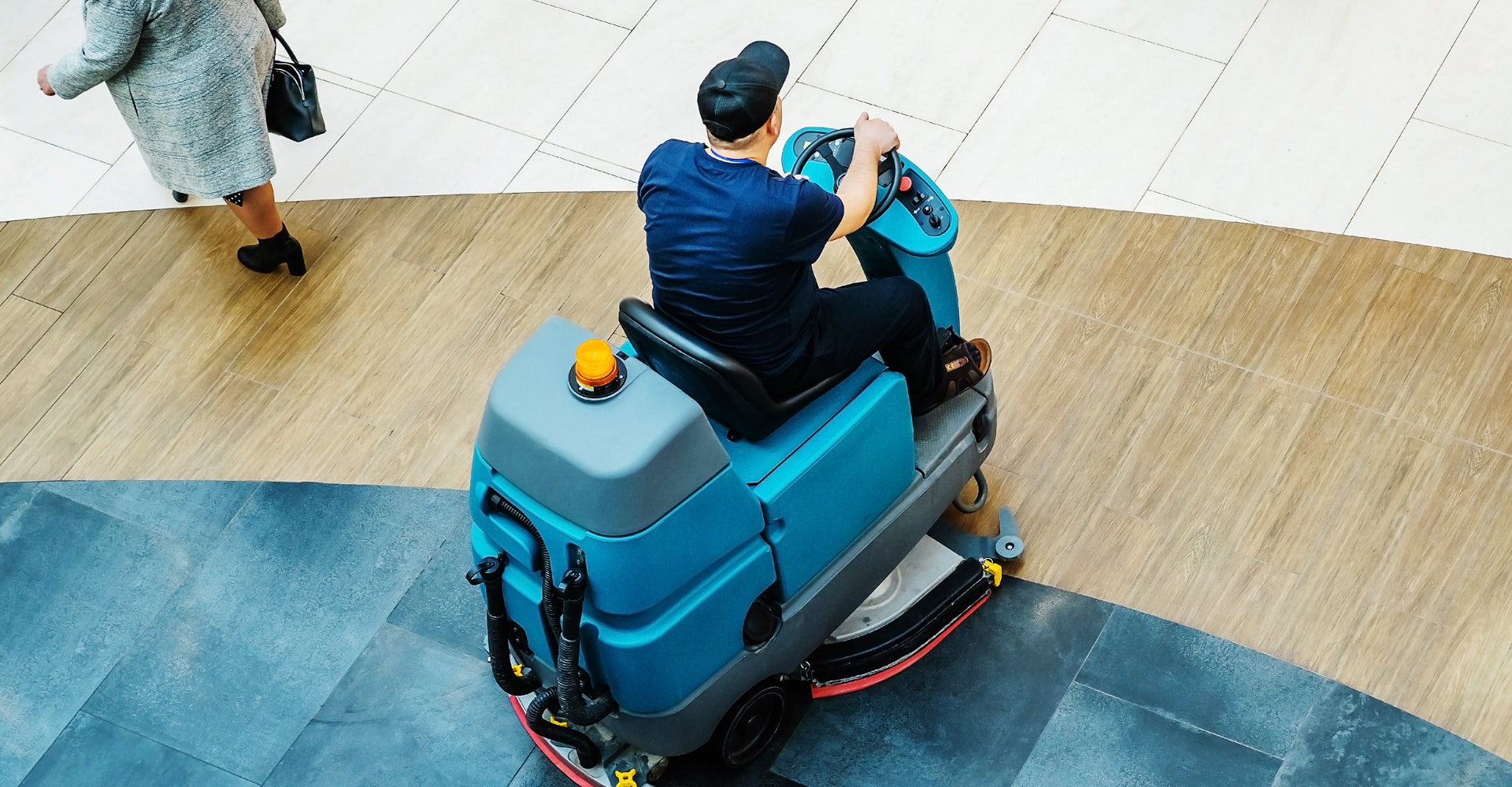Agent de nettoyage sur autolaveuse bleue professionnelle, nettoyant un sol carrelé dans un espace public moderne. Une femme marche à côté.