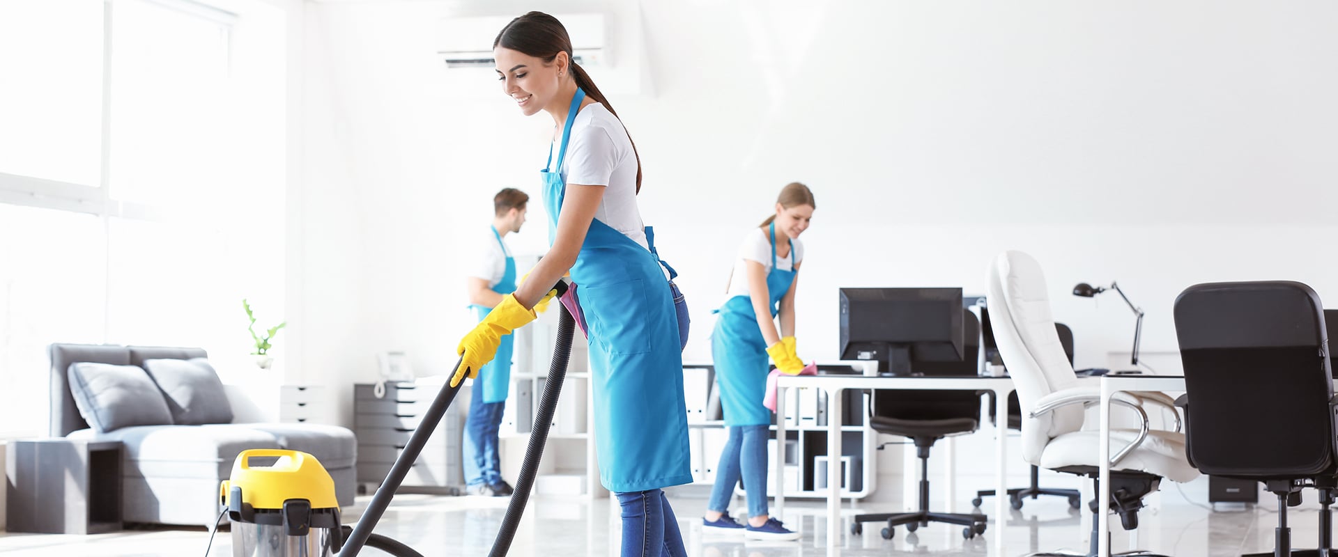 Équipe de nettoyage professionnelle souriante aspirant et essuyant un bureau lumineux. Agents en uniforme bleu et gants jaunes travaillent.